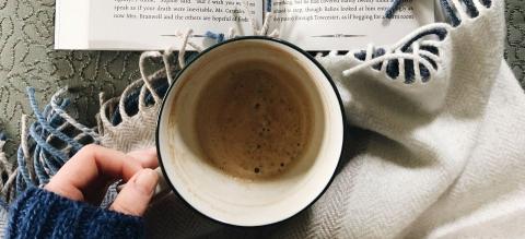 A hand holding a half-full coffee mug on top of a fringed blanket. A book is open above the mug.