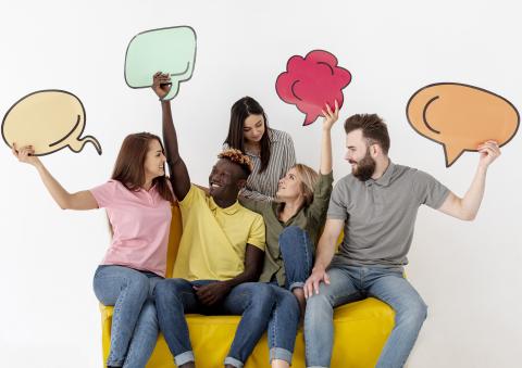 Group of five people sitting on a yellow couch and looking at each other and holding pictures of talking, or speech, bubbles