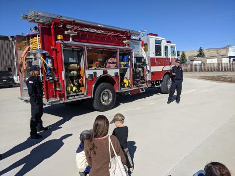 A mother and her children are listening to two firefighters give a show-and-tell in front of a fire truck.