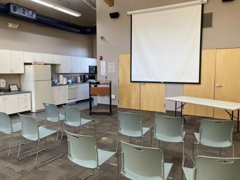 Large room with fridge, stove, cabinets, pull down screen, projector, and chairs.