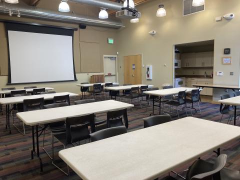 A large room with 6 tables, each with 4 chairs. A projection screen and small kitchen are along walls