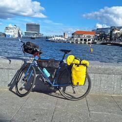 A blue bicycle loaded with black and yellow travel gear. Background is a river front with tall and small buildings.