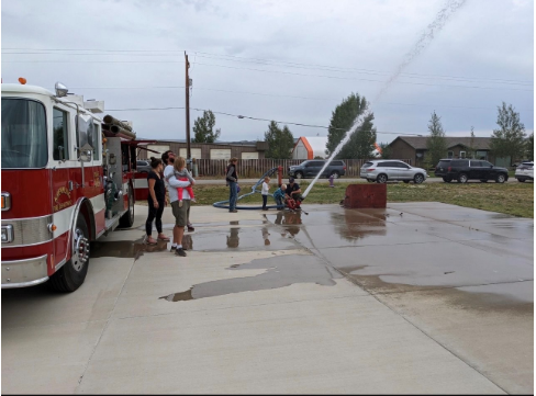 A firefighter and child are shooting water from a hose while people are watching. A fire truck is behind them.