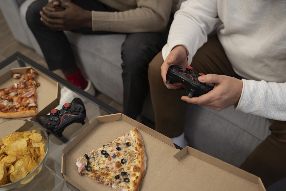 Two people sitting on a couch. The zoom in shows hands with video game controllers. Pizza slices and chips are on a table.