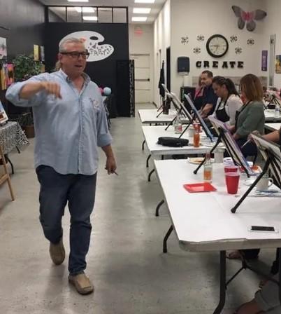 Man standing next to a long, white table where people are sitting and painting on canvas